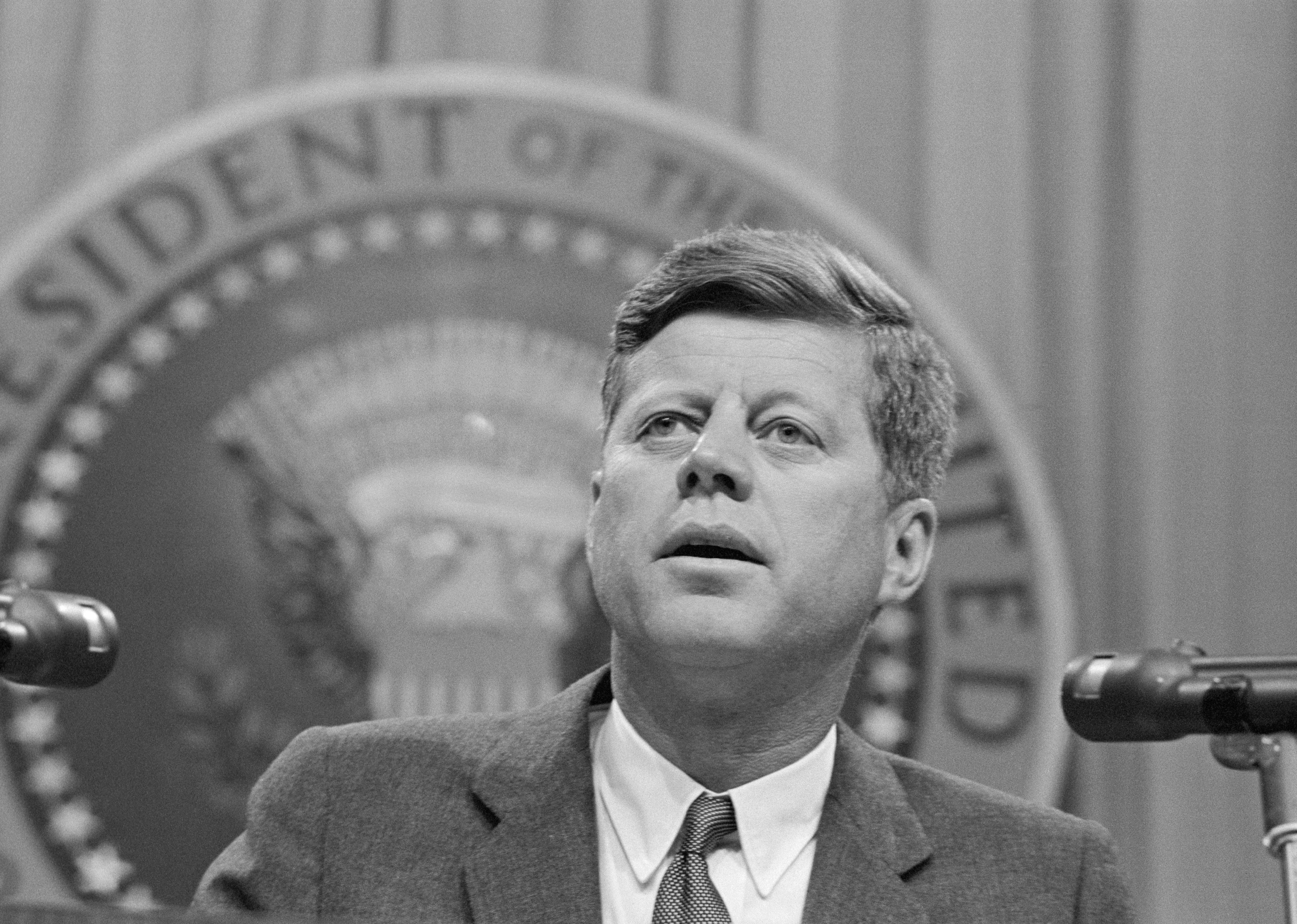 John F. Kennedy speaking in front of the presidential seal.