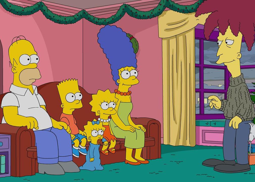 The Simpsons family on the couch while a man with wild hair stands in front of them.