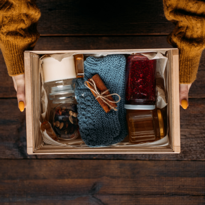 Two hands holding a wooden crate filled with cozy goods 