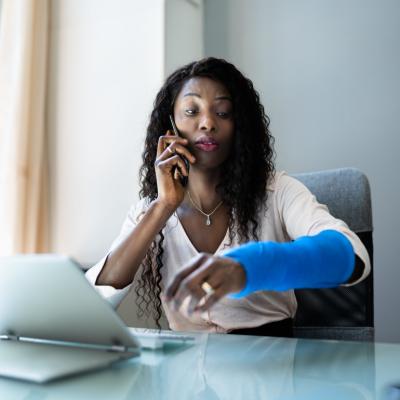 A woman at a desk on the phone with a blue cast on her arm.