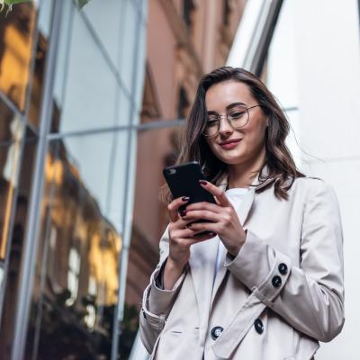 Smiling woman wearing sunglasses walks down a city street and uses her phone.