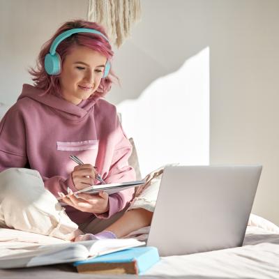 Student with pink hair with headphones takes notes while watching online webinar.
