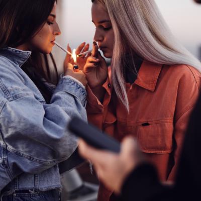 Two teenage girls standing outdoors and lighting cigarettes.