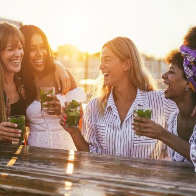 A group of women laughing and drinking cocktails outside as the sun is setting. 