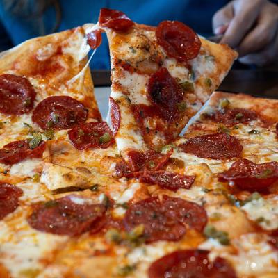 Slice of appetizing pepperoni pizza with melting cheese is being up and picked up from whole pizza by a person off screen.