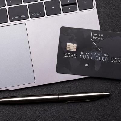 Black premium bank card lays on a silver macbook laptop computer next to the trackpad, with a shiny fancy pen on the table.