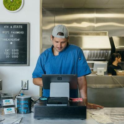 Person wearing a blue shirt entering items into a tablet cash register behind the counter with a kitchen and sign int he background.