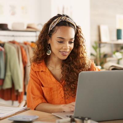 Black woman using laptop in clothing store working on business with rolling rack of clothes behind her.
