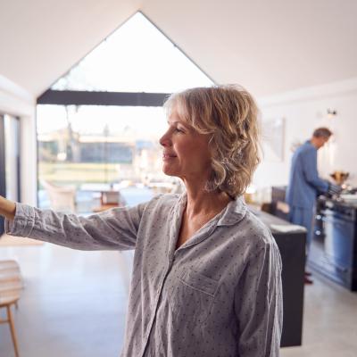Middle-aged woman wearing pajamas adjusts modern thermostat in a bright house with a vaulted ceiling while a person wearing blue pajamas is seen in the kitchen counter in the background.