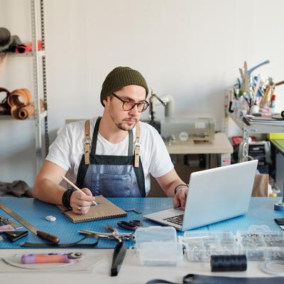 A leather goods craftsman using a laptop and notebook to start his production process.