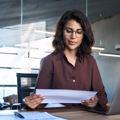 Focused young businesswoman reading financial report document and using a laptop in the workplace.