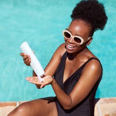 Black woman wearing black suit and white-framed sunglasses applies sunscreen sitting by pool in summer sun