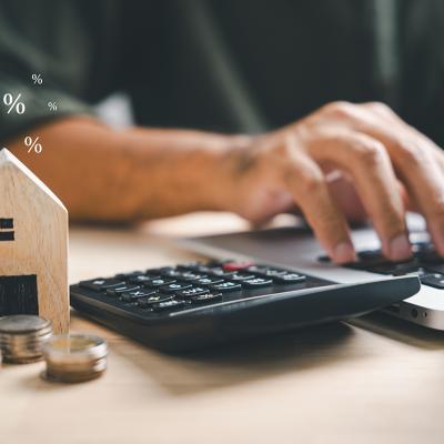 A person using a laptop and calculator with a wooden figure of a house and coins beside him.