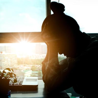 Silhouete of person sitting at desk in front of laptop, with head in hands backlit by sunset shining through office window in background.