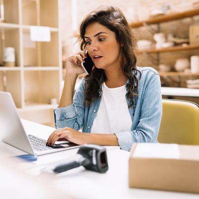 A businesswoman speaking on the phone while using a laptop in her small business production area.