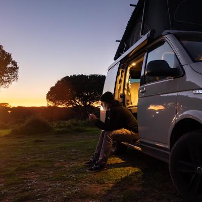Silhouetted digital nomad sitting in his white transporter camping van, backlit by the setting sun near the California coast.