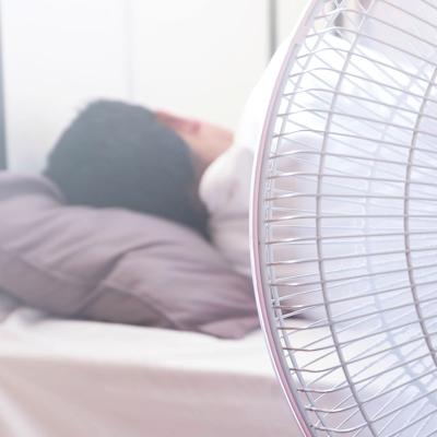 A white electric fan in the foreground in front of a person sleeping in bed, facing away.
