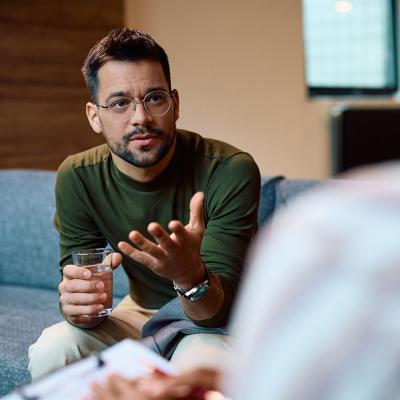 Person wearing long sleeve forest green top and glasses sits on a couch holding a glass of water and gesturing to a person with their back to their camera.