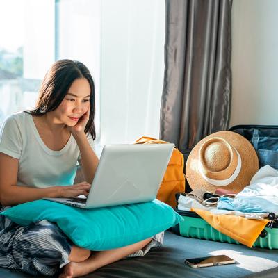Young woman using a laptop beside an unpacked suitcase preparing for a trip.