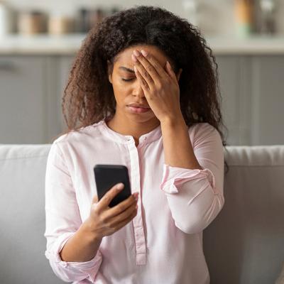 A photo capturing the tension of a young African American woman sitting on her couch, visibly overwhelmed as she looks at her smartphone in a neatly arranged living room.