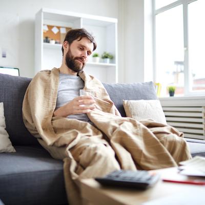 Sick man wrapped into blanket sitting on sofa in front of table with papers while staying at home.