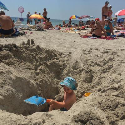 Child wearing a blue hat digging a giant hole in sand at the beach that is higher than him on both sides.