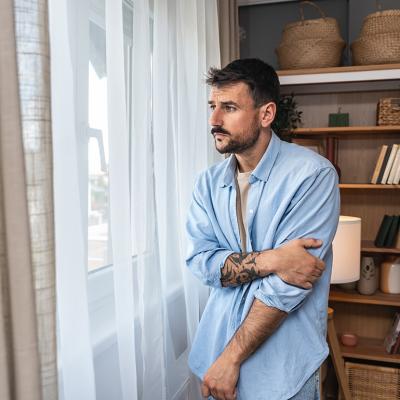 Young sad and depressed anxious looking man wearing a blue button up shirt looking through white curtains out the window.
