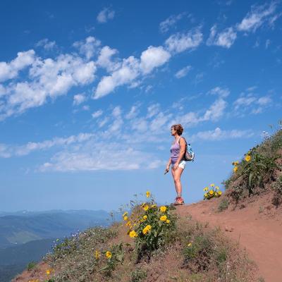 Woman looks out toward view on dirt mountain trail with yellow flowers, hiking in summer with sky and clouds in background near Portland, Oregon.