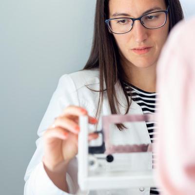 Serious looking woman in doctor's coat adjusting scale to weigh patient, whose back is to us in the foreground.