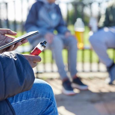 Teenager holding a vape while scrolling on the phone at the park.