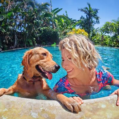A medium-sized brown dog and blond woman look at each other happily in the pool, with their hands and paws holding onto the edge of the pool.