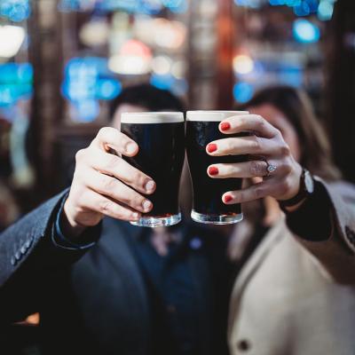 Two people holding up a pint each of stout beers to cheers, which obscures their faces from the camera in a bar.