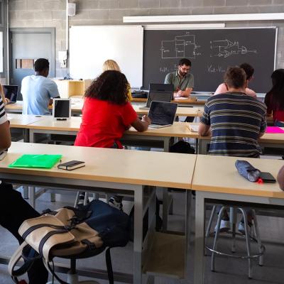 View from back of classroom of multiracial high school students in class using laptops while teacher marks exams.