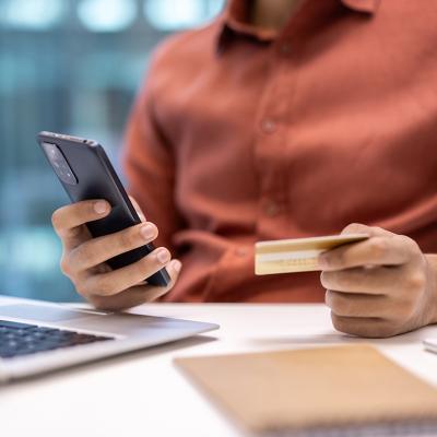 Businessperson holding a credit card while using a phone and laptop at an office desk.