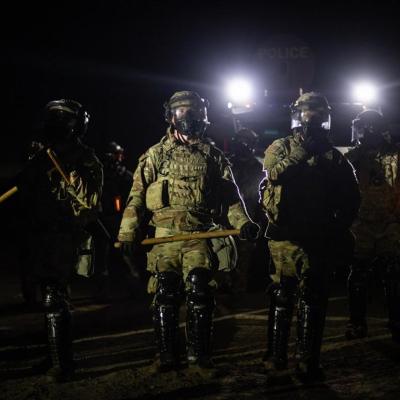 Nighttime view of a line of federal immigration agents in riot gear and face shields facing the camera near Glass House Farms facility in Camarillo, with police spotlights in background.