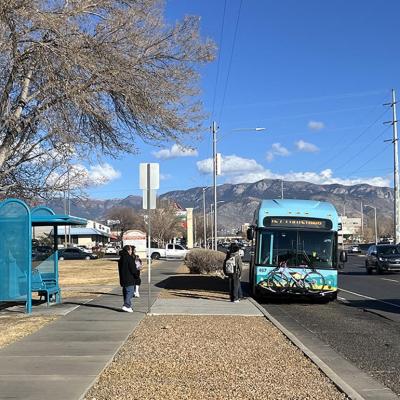 Bus riders board a blue bus in Albuquerque, next to a turquoise metal bus stop and with mountains and cloud in distance.