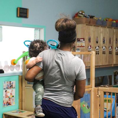 A child care teacher carrying an infant in a child care room.