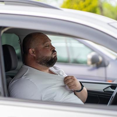 A bald driver with closed eyes seen through the passenger side window pulls at the collar of a white t-shirt and exhales.