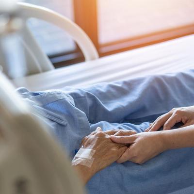 Elderly senior patient in hospital bed, whose face and torse are not visible, has hand held by younger caretaker.