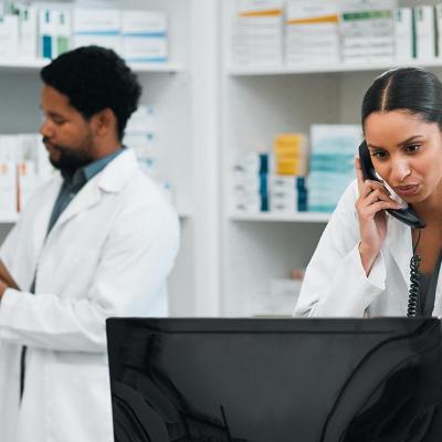 Two pharmacy employees work behind the counter in a pharmacy, one on the phone and looking at a computer and the other writes on a clipboard.