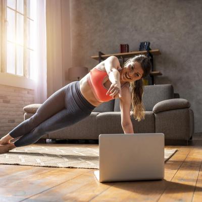 Woman doing a side plank pose on a mat on a wooden floor, following a video on a laptop, with sunlight coming in through large window in the background. 
