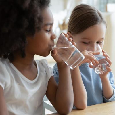 Two children, one Black with an afro and one light skinned child with long hair tied back sit at a table drinking glasses of water.