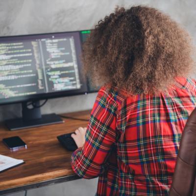 Rear view of a woman with curly hair working at a computer desk displaying code in a casual office setup wearing a checked shirt.