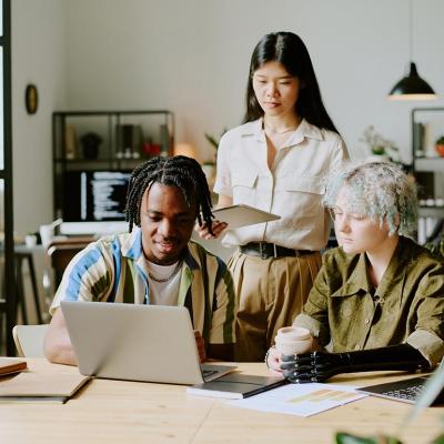 Three diverse young adults gathered around a laptop in a modern office setting.