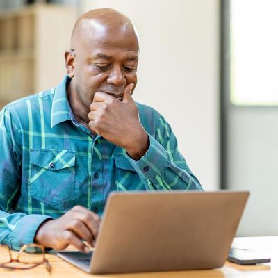 Black bald man wearing teal button up shirt using laptop computer and thinking about question in office.