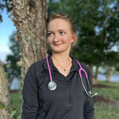 Michaela Bonner, wearing a black zip-up top with a pink stethoscope around her neck, poses for a portrait in front of some trees at local hospital in Virginia Beach, VA.