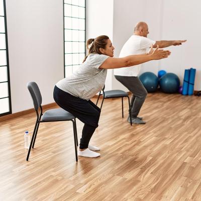 Two people each squatting toward a chair with their arms out in front of them, in a yoga studio type room.