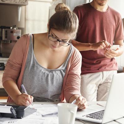 Woman doing calculations at kitchen table with laptop open and pen and paper while man with face not visible in the background is on his phone.