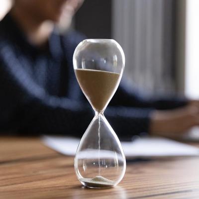 Close up view of hourglass stand on wooden office table measuring time, person working on laptop in the background blurred.