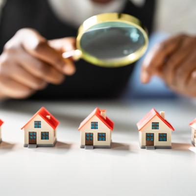 Five miniature houses with red roofs lined up on table, with magnifying glass held above them by a person symbolizing home and neighborhood appraisals.